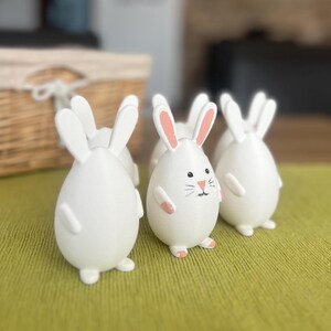 Könnte beinhalten: Fünf weiße Keramik-Hasenfiguren mit rosa Nasen und Ohren. Die Hasen sind in einer Reihe auf einer grünen Oberfläche angeordnet.