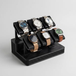 Op de afbeelding: Een zwarte horlogestandaard met zeven polshorloges. De horloges hebben verschillende band- en wijzerplaatkleuren, waaronder zilver, goud en bruin. De standaard is gemaakt van zwart plastic en heeft een geribbeld ontwerp op de basis.