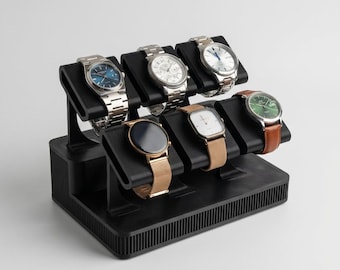 May include: A black watch display stand holding seven wristwatches. The watches have various band and face colors, including silver, gold, and brown. The stand is made of black plastic and has a ribbed design on the base.