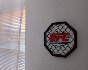 May include: A black octagon-shaped wall decor with a red UFC logo and a black wire mesh design. The text "ULTIMATE FIGHTING CHAMPIONSHIP" is written in white letters below the logo.