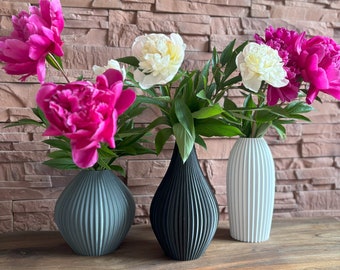 May include: Three ceramic vases with a textured, ribbed design. The vases are in shades of gray, black, and white. The vases are filled with pink and white peonies.