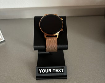 May include: A gold smartwatch with a black face and a metal band is displayed on a black stand with the text "YOUR TEXT" on a label.