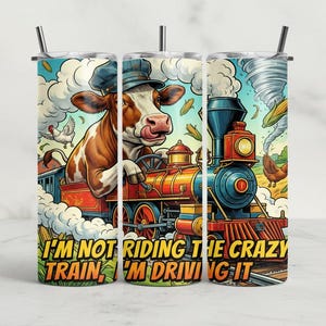 May include: A stainless steel tumbler with a whimsical illustration of a cow driving a train. The design features a cow wearing a conductor's hat, a train, chickens, corn, and the text "I'M NOT RIDING THE CRAZY TRAIN, I'M DRIVING IT."