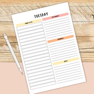 May include: A white paper planner for Tuesday with sections for things to do, top priority, reminders, and notes. The planner has a light pink and yellow color scheme. A white pencil with a pink eraser rests on the planner.