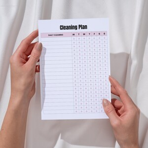 May include: A white Cleaning Plan notepad with a pink border is held by hands. The notepad has a grid for daily cleaning tasks, with the title "Cleaning Plan" at the top. The days of the week are listed across the top.