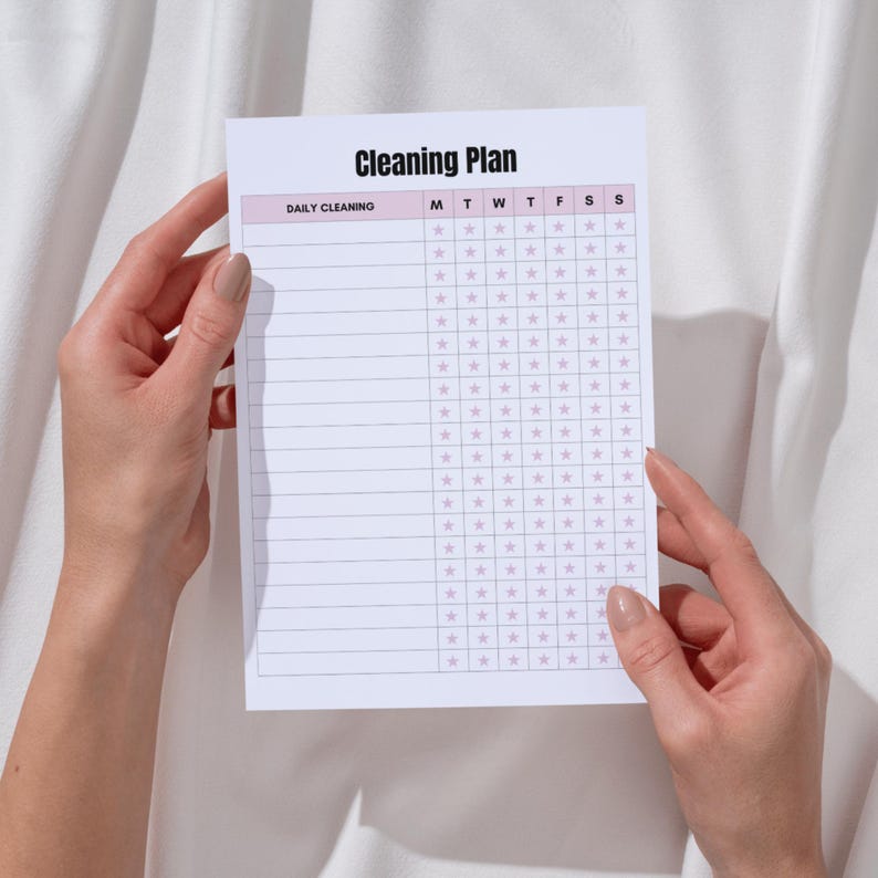 May include: A white Cleaning Plan notepad with a pink border is held by hands. The notepad has a grid for daily cleaning tasks, with the title "Cleaning Plan" at the top. The days of the week are listed across the top.