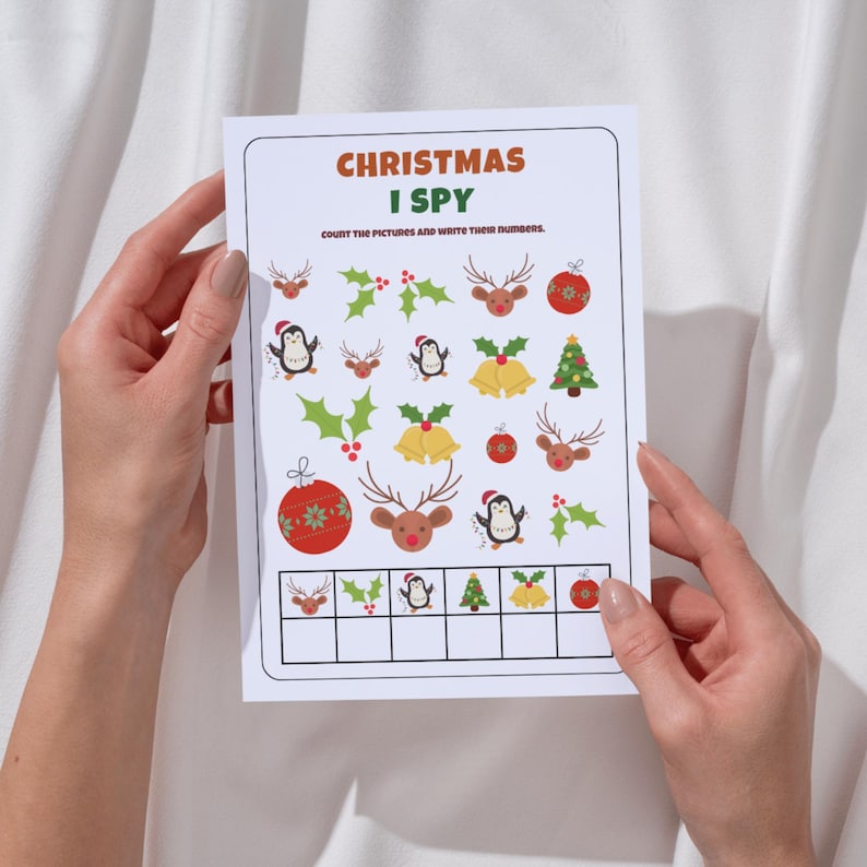 May include: A Christmas I Spy game sheet held by hands. The game sheet features colorful illustrations of Christmas ornaments, reindeer, penguins, holly, and bells. The text "CHRISTMAS I SPY" is at the top, with instructions below.
