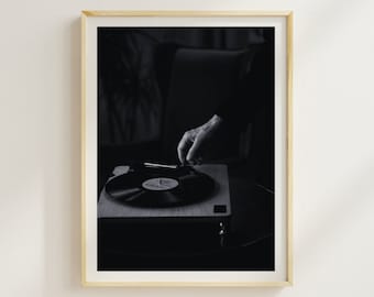 May include: A black and white photo of a hand placing a vinyl record on a turntable. The turntable is on a wooden surface and the hand is reaching from the left side of the image.