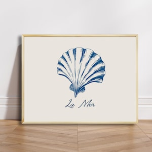 May include: A framed print with a blue seashell design on a cream background. The words "La Mer" are written in a script font below the seashell. The frame is a light wood colour.
