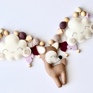 May include: A baby mobile featuring a brown deer, two white clouds with happy faces, wooden beads, and purple felt leaves. The mobile has two purple plastic clips.