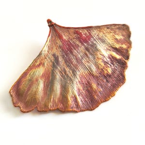 Könnte beinhalten: Ein fächerförmiges Ginkgo-Blatt-Ornament mit metallischem Finish in gemischten Kupfer-, Bronze-, Gold- und Dunkelrottönen. Die Oberfläche zeigt feine, strahlenförmige Adern, die vom Stiel bis zum gewellten, geschwungenen Außenrand verlaufen.