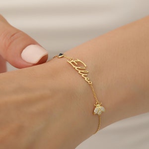 Könnte beinhalten: Goldkette Armband mit dem Namen "Ellie" in Kursivschrift und einem kleinen, funkelnden Bienenanhänger. Das Armband hat zwei kleine schwarze Edelsteine.