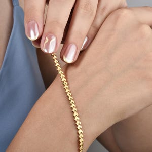 May include: A gold bracelet featuring a repeating heart pattern. The bracelet is worn on a wrist, with the hand and arm of a person visible. The person is wearing a blue top. The nails are painted with a light pink and white colour.