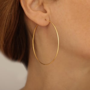 Puede incluir: Primer plano de un pendiente de aro dorado. El pendiente es un diseño circular grande y delgado. El pendiente es de color dorado brillante y brillante. El pendiente se lleva en la oreja de una persona con piel clara y pelo corto.