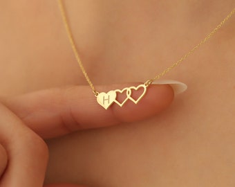 May include: A gold necklace with three heart-shaped pendants. The first heart is solid and engraved with the letter "H", the others are open. The chain is fine. The necklace is shown against a blurred background.