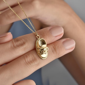 May include: A gold-colored pendant necklace featuring a small shoe charm. The charm has a bow detail and a small gem. The necklace is on a delicate chain. The background is a blurred view of a person's hand.