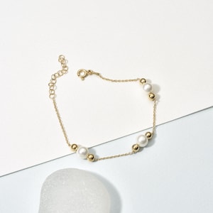 May include: A delicate gold bracelet featuring a chain with alternating white pearls and small gold beads. The bracelet has a lobster clasp closure and an adjustable chain. The jewelry is displayed on a white and light blue surface.