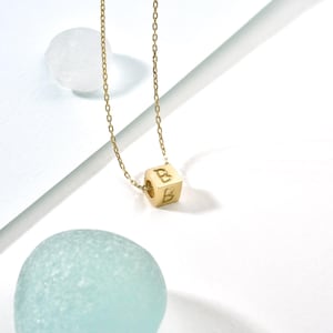Op de afbeelding: Een gouden ketting met een gouden kubus hanger met de letter 'B' aan elke kant.