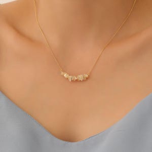 Puede incluir: Un collar de cadena dorada con una fila delicada de pequeñas piedras irregulares de color blanco roto.