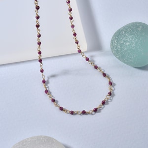 Può includere: Una delicata collana con catena dorata con piccole perline rotonde rosso scuro. Le perline sono uniformemente distanziate lungo la catena dorata, creando un design semplice ma elegante. La collana è esposta su una superficie azzurra.