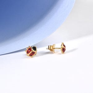 May include: A pair of gold-colored stud earrings featuring a ladybug design. Each earring has a red body with black spots and a black head. The earrings are set against a white and blue background.