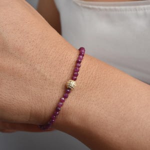 May include: A fine bracelet with small, faceted, deep red beads. A gold-coloured, diamond-studded sphere is at the centre. The bracelet is worn on a wrist against a neutral background.