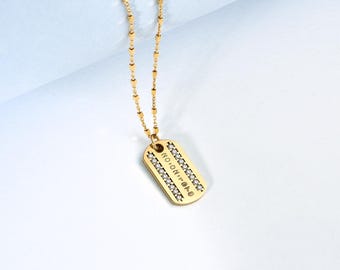 May include: Gold-toned dog tag pendant necklace with a delicate chain. The rectangular tag is embellished with small, sparkling stones and features engraved text. The necklace is displayed against a white and light blue background.
