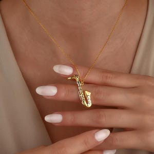 Pode incluir: Um colar com pingente de saxofone em tom dourado, com acabamento polido e pequenas pedras claras e brilhantes incrustadas ao longo do corpo do instrumento. O pingente está suspenso por uma corrente fina e delicada de cor dourada.