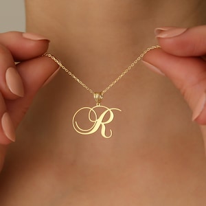 Puede incluir: Un collar dorado con un colgante con la letra "R" en cursiva. El colgante cuelga de una delicada cadena. El collar está sostenido por las manos de una persona.