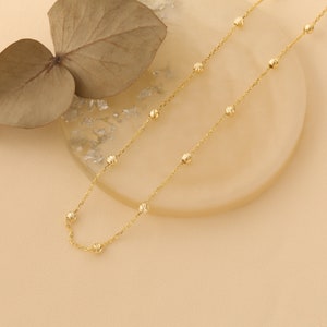 May include: A delicate gold chain necklace with small, round beads. The chain is laying on a light brown surface.
