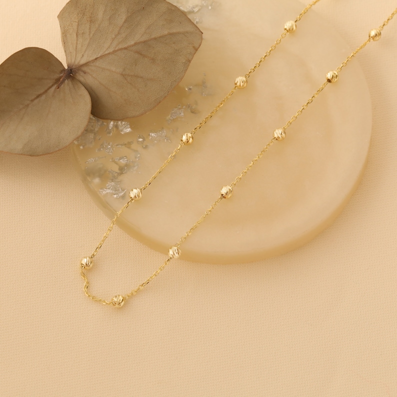 May include: A delicate gold chain necklace with small, round beads. The chain is laying on a light brown surface.