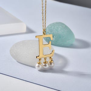 May include: A gold-coloured necklace featuring the letter "E" pendant. The pendant has three small, dangling pearls. The necklace is displayed on a white and blue background, with a light blue stone in the background.