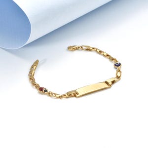 May include: Gold bracelet with a rectangular nameplate. The bracelet has a chain design, a small red and black charm, and a blue and white eye charm. The bracelet is shown on a white surface with a blue backdrop.