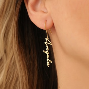 May include: Gold-colored, personalized dangle earrings. The earrings feature a curved hook design and the name "Angela" written in a cursive font. The earrings are shown worn on a person.