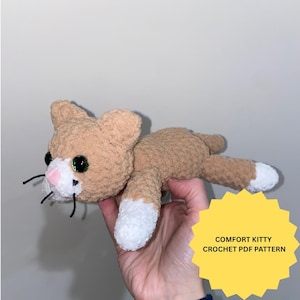 May include: A tan and white crocheted cat plush toy with dark green eyes, a pink nose, and black whiskers. A yellow star-shaped graphic overlay in the bottom right corner contains the black text COMFORT KITTY CROCHET PDF PATTERN.