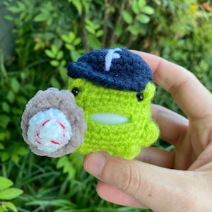 Può includere: Una creatura verde fatta a mano all'uncinetto con un berretto da baseball blu navy con una "F" bianca e che tiene un guanto da baseball con una palla bianca e rossa. La creatura ha occhi neri e una bocca verde chiaro.