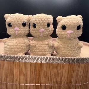 Może przedstawiać: Trzy beżowe figurki kota na szydełku z czarnymi oczami i różowymi nosami i pyszczkami. Koty siedzą w okrągłym, jasnobrązowym bambusowym koszu z białą podszewką z tkaniny. Tło jest ciemne.