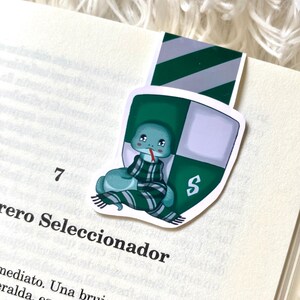 May include: A green and silver bookmark featuring a cartoon snake wearing a green and white striped scarf. The snake is curled up and has a sad expression. The bookmark has the letter "S" on it.
