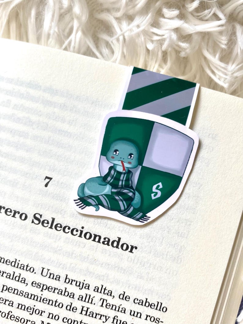 May include: A green and silver bookmark featuring a cartoon snake wearing a green and white striped scarf. The snake is curled up and has a sad expression. The bookmark has the letter "S" on it.