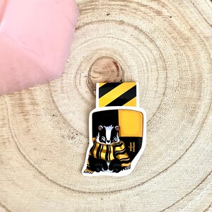 May include: A black and yellow badger wearing a black and yellow scarf with a black and yellow H on a white background. The badger is a mascot for a fictional house in a book series.
