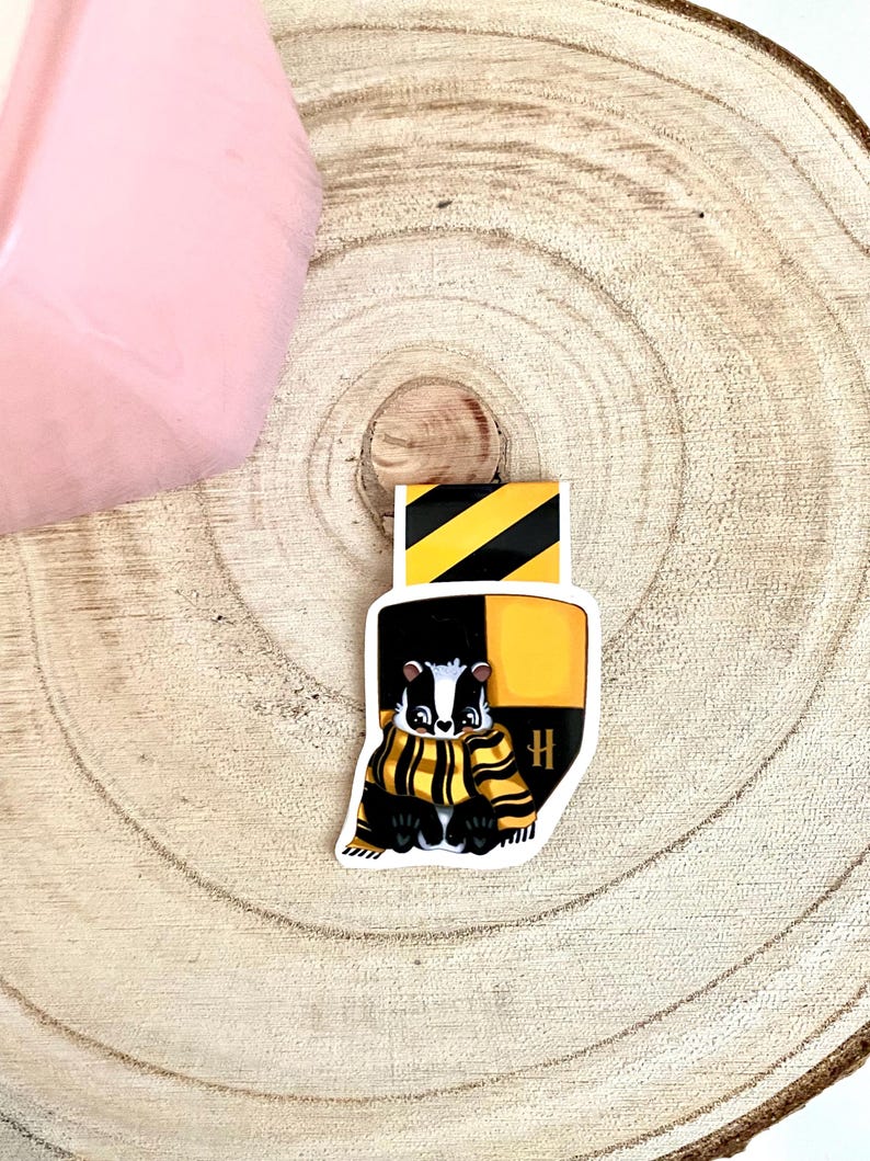 May include: A black and yellow badger wearing a black and yellow scarf with a black and yellow H on a white background. The badger is a mascot for a fictional house in a book series.