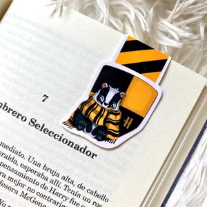May include: A yellow and black striped bookmark with a cartoon badger wearing a yellow and black scarf. The bookmark has a large "H" on it.