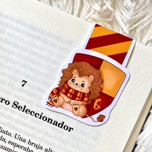 May include: A Gryffindor house bookmark featuring a cartoon lion wearing a red and gold scarf. The bookmark is shaped like a shield with the Gryffindor house crest and the number "6" on it.
