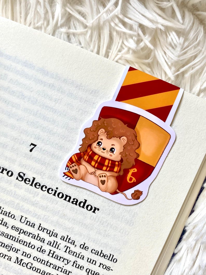 May include: A Gryffindor house bookmark featuring a cartoon lion wearing a red and gold scarf. The bookmark is shaped like a shield with the Gryffindor house crest and the number "6" on it.
