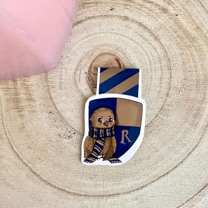 May include: A brown and white owl wearing a blue and gold scarf with the letter "R" on a shield. The owl is a bookmark.