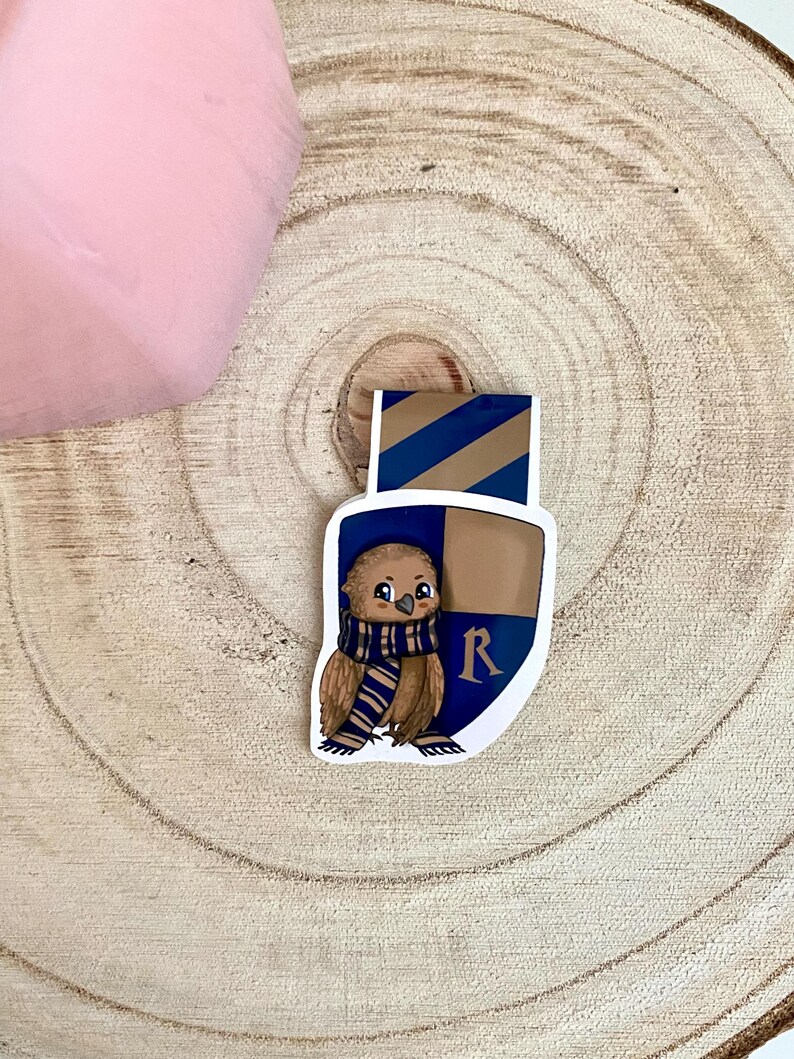 May include: A brown and white owl wearing a blue and gold scarf with the letter "R" on a shield. The owl is a bookmark.