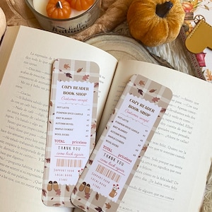 Könnte beinhalten: Ein herbstliches Bild mit einer Kerze, die mit drei orangefarbenen Kürbisdekorationen, einem kleinen Samtkürbis und zwei Lesezeichen verziert ist. Die Lesezeichen listen Artikel des Cozy Reader Book Shop auf. Im Hintergrund ist ein offenes Buch zu sehen.