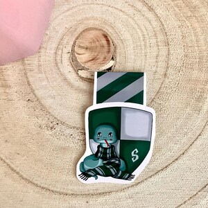 May include: A green and white bookmark with a cartoon snake wearing a green and white scarf. The bookmark has the letter "S" on it.