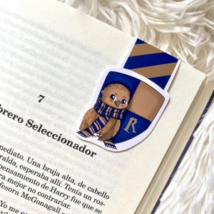 May include: A bookmark featuring a cartoon illustration of a brown owl wearing a blue and gold scarf. The owl is perched on a shield with the letter "R" in the center. The shield is blue and gold with a diagonal stripe pattern.