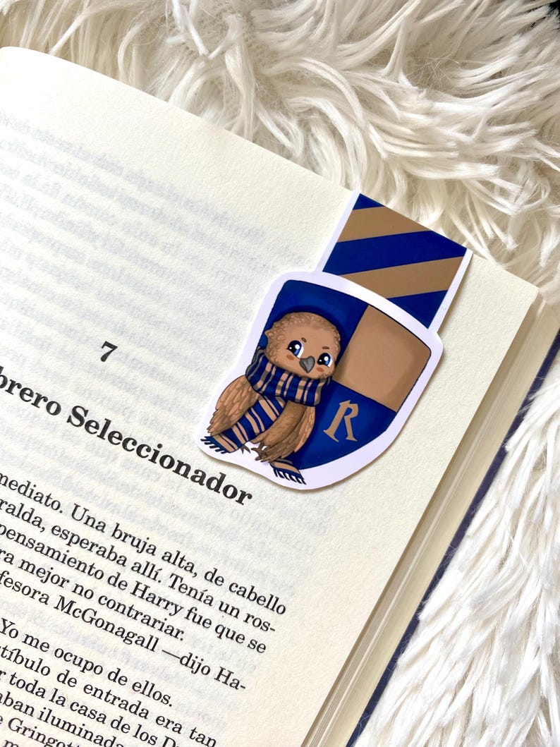 May include: A bookmark featuring a cartoon illustration of a brown owl wearing a blue and gold scarf. The owl is perched on a shield with the letter "R" in the center. The shield is blue and gold with a diagonal stripe pattern.